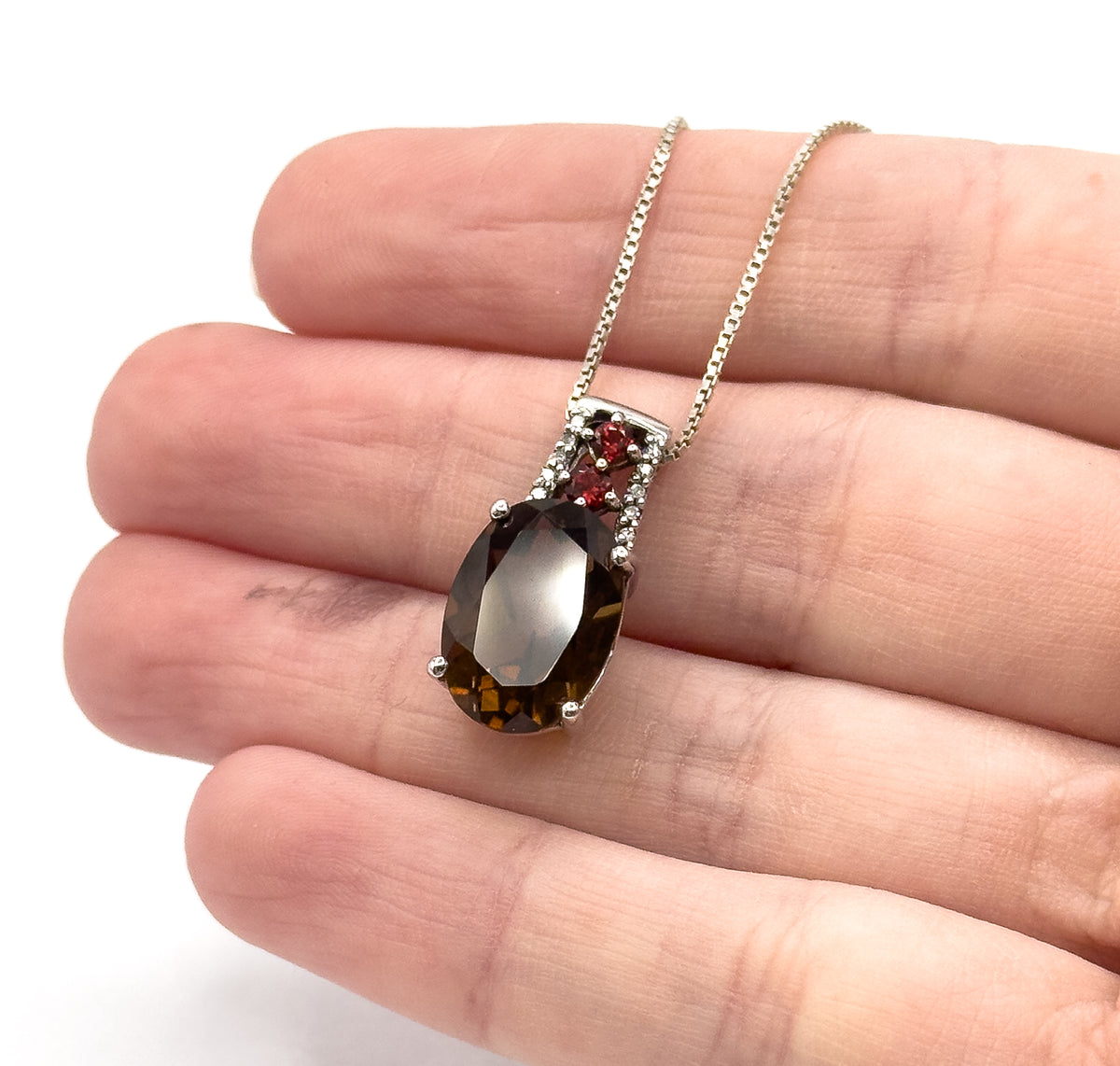 The pendant shown resting across the fingers of a hand, giving a sense of its small scale, with the oval quartz, garnets and diamonds facing forward and the box chain rising behind.