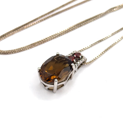 The whole necklace laid out on white, the box chain looping wide above the pendant whose amber-brown oval quartz sits in prongs beneath a bar of small garnets and clear stones.
