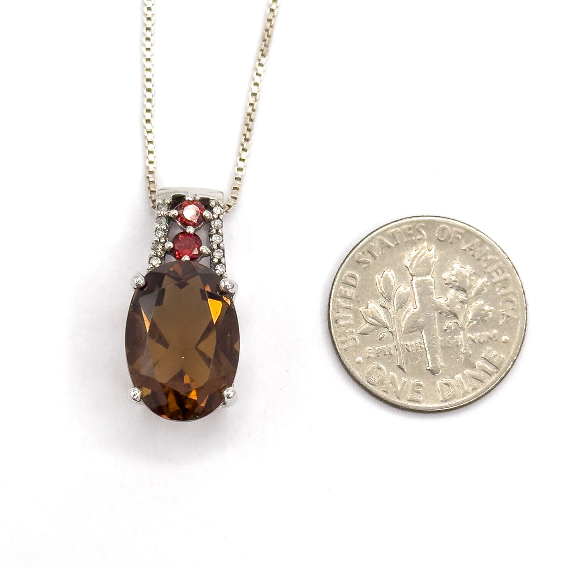 The pendant lies flat beside a dime for scale, the oval quartz standing roughly as tall as the coin is wide, with the garnets and tiny clear stones above it.