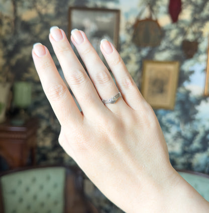 Mid-Century platinum V-shaped band set with three full-cut diamonds at the point, shown on a ring finger of a raised hand against a soft blue-green papered room.