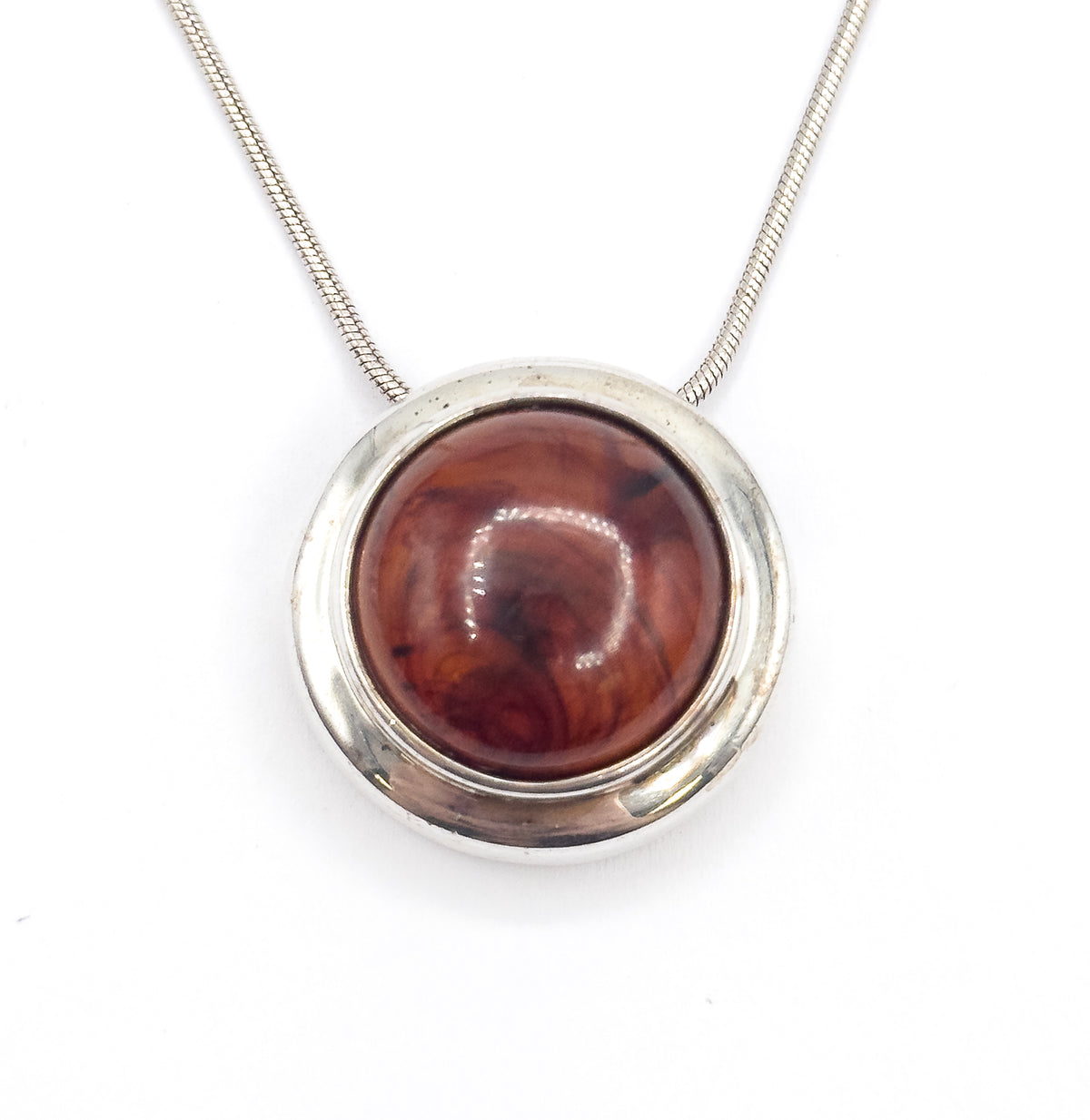 Straight-on front view of the round pendant lying flat on white, the amber-coloured resin cabochon and its dark swirls filling the wide sterling silver bezel, with the snake chain rising in a V above.