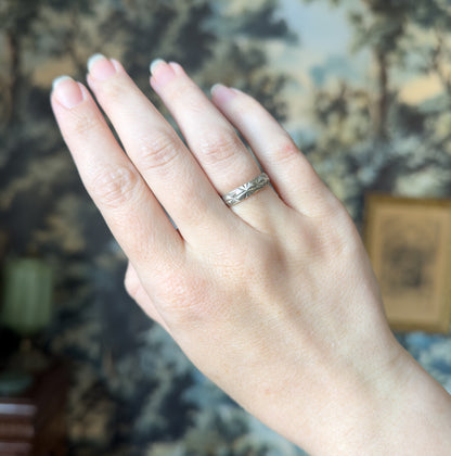 Shown on the ring finger of a raised hand held against a softly blurred scenic backdrop, the band's narrow faceted shank giving a clear sense of its modest width.
