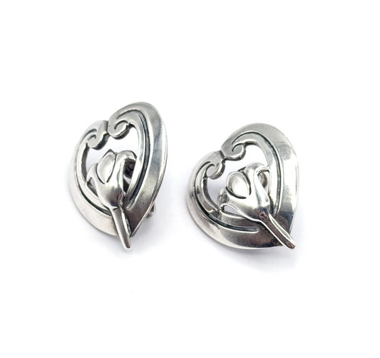 The pair angled three-quarter on white, tilted to show the domed curve and thickness of each heart and the screw back fitting sitting behind the left one.