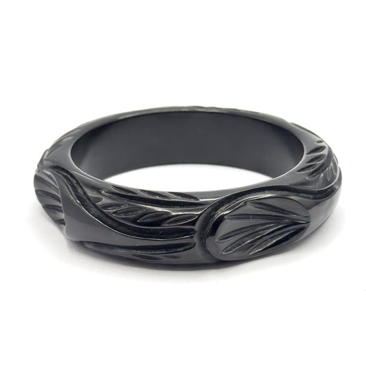 Mid-century hand carved black Bakelite bangle, shown at a slight angle on a white background so the wide leaf-and-scroll carving around the outside and the smooth interior are both visible.