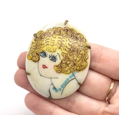 The oval plaque rests on an open palm, filling most of the fingers, with six base metal prongs gripping its edge and the painted flapper's blue beads visible at her neckline.