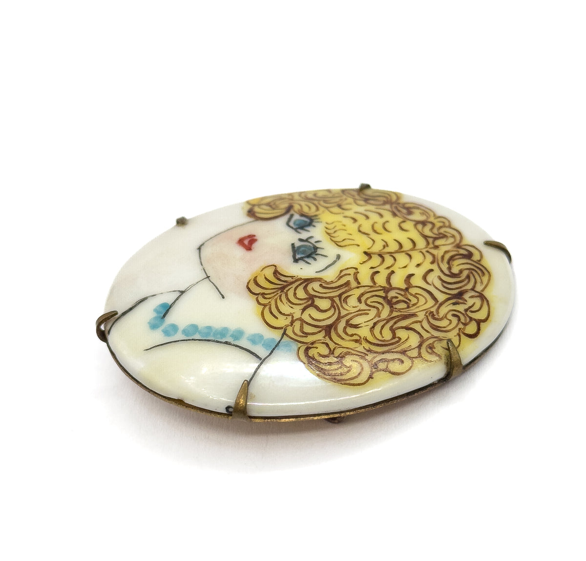 A low three-quarter view from the side, the oval tilted so the painted flapper's curls and blue beads face the camera and the prongs are seen gripping the plaque's thickness.