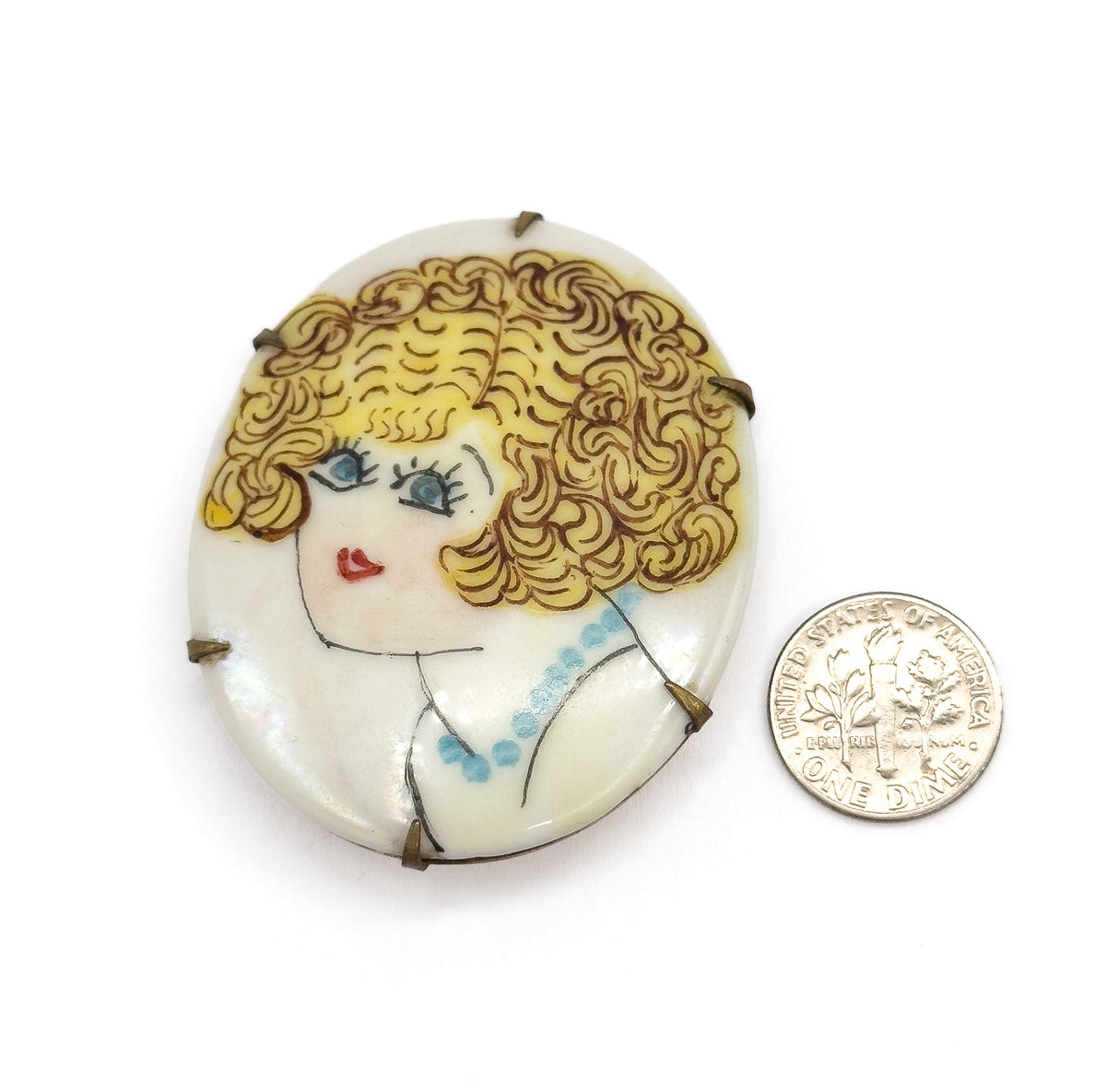 Art Deco hand-painted porcelain flapper brooch in base metal, shown face-up beside a dime for scale, the oval plaque held by small metal prongs around its edge.