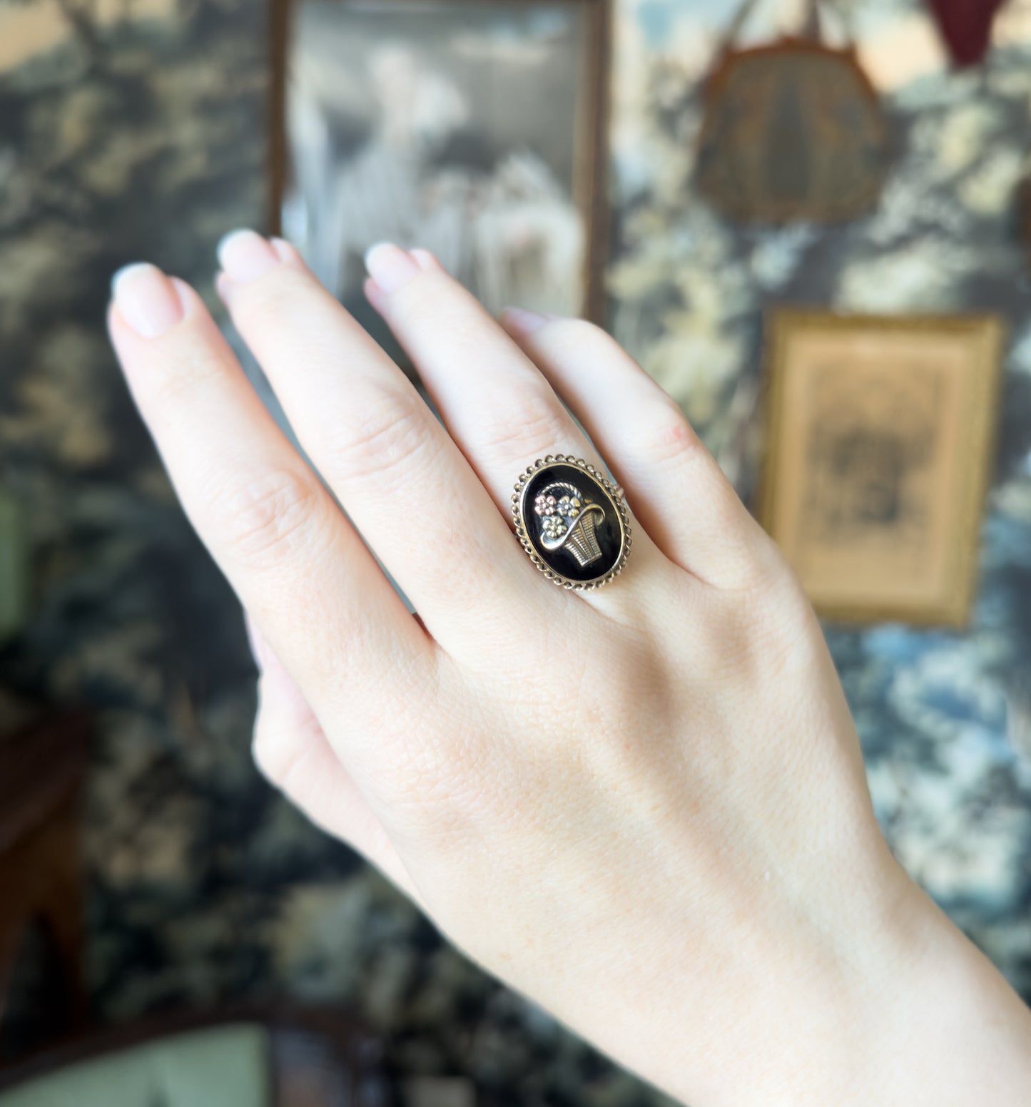 Shown on the ring finger of a raised hand held sideways, the oval face reading as a small dark oval against the skin, with framed pictures and patterned wallpaper blurred behind.