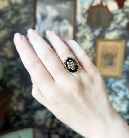 Shown on the ring finger of a raised hand held sideways, the oval face reading as a small dark oval against the skin, with framed pictures and patterned wallpaper blurred behind.