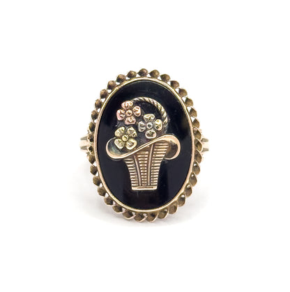 Straight-on view of the ring face filling the frame, the oval black onyx ringed by its twisted rope bezel with the applied tri-color basket and five flowers centred against the stone.