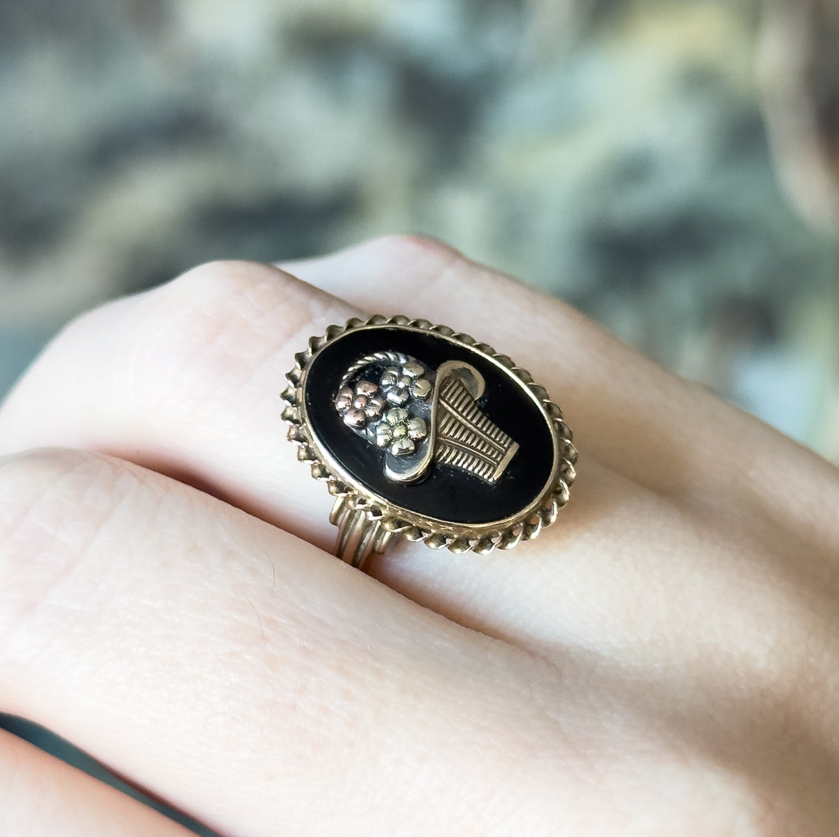 Shown on a finger at a slight tilt, the oval face spanning nearly the full width of the finger, with the rope border and applied flowers catching the light against a soft green blurred background.