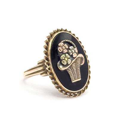 Mid-century black onyx flower basket ring in tri-color gold, shown at a three-quarter angle on white, with the oval bezel and rope border, applied basket and flowers, and grooved shank in view.
