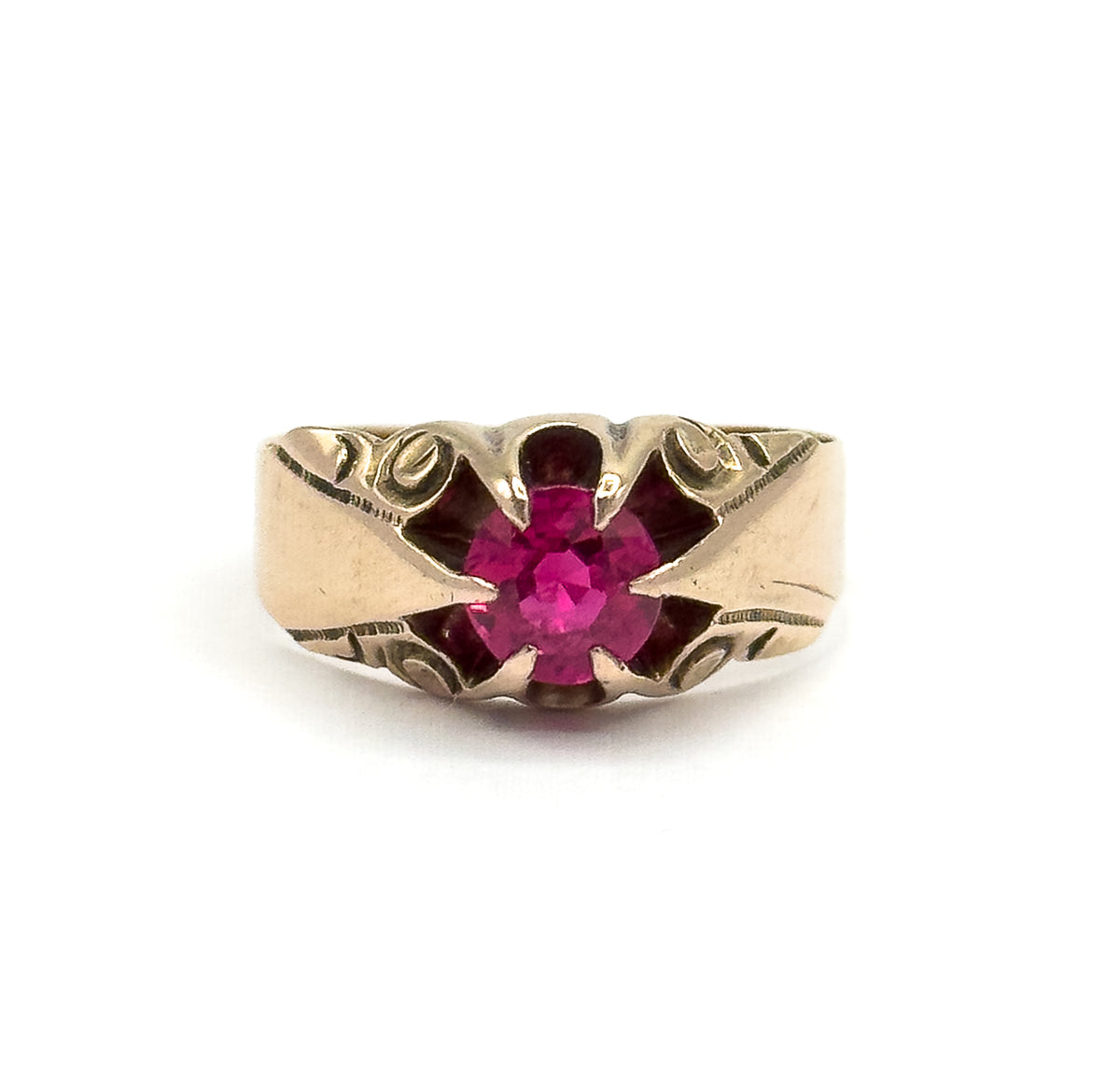 Full frontal view of the ring standing upright, the round pinkish red stone centred in its four prongs with engraved swirls on both shoulders and the wide band narrowing below.