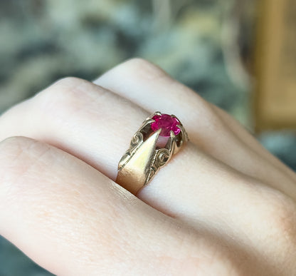 Close side view on the finger, the raised four-prong head lifting the pinkish red stone above swirl-engraved shoulders that taper into the wide polished shank.