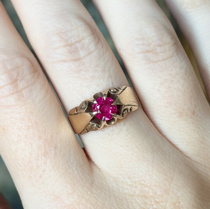 A wide-band 10K yellow gold ring with a round synthetic ruby in a raised four-prong head, shown on a finger with engraved swirls at the shoulders.