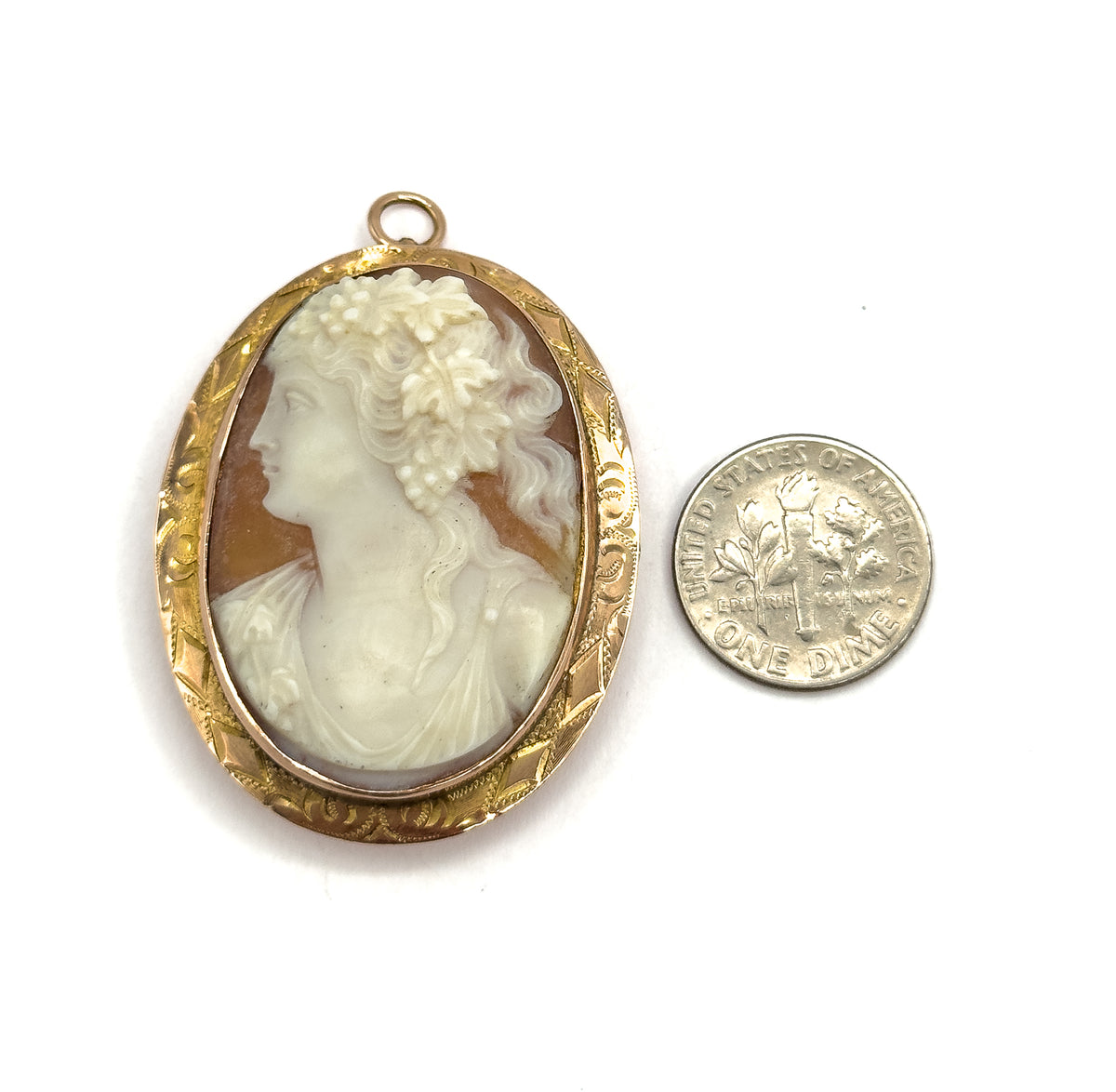 Front view laid flat beside a dime, the oval cameo and its engraved gold frame standing about twice the coin's width, with the small round bail at the top.