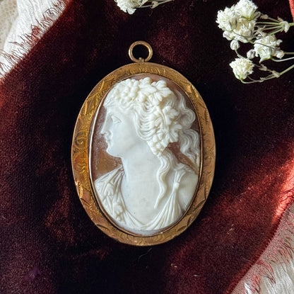 Close-up on deep red velvet beside sprigs of tiny white flowers, the carved curls and grape leaves of her hair crisply detailed above the hand-engraved scrollwork of the gold bezel.
