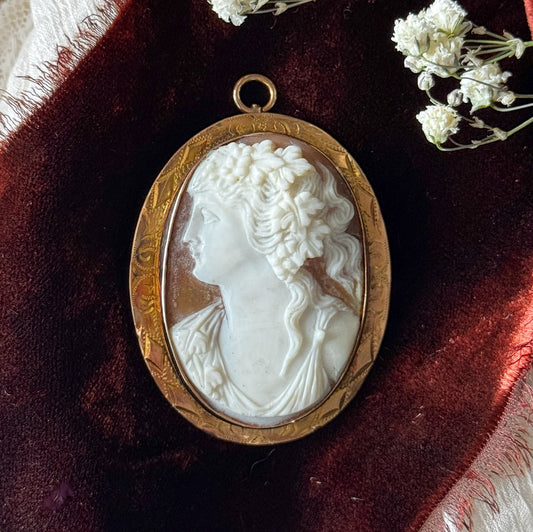 Close-up on deep red velvet beside sprigs of tiny white flowers, the carved curls and grape leaves of her hair crisply detailed above the hand-engraved scrollwork of the gold bezel.