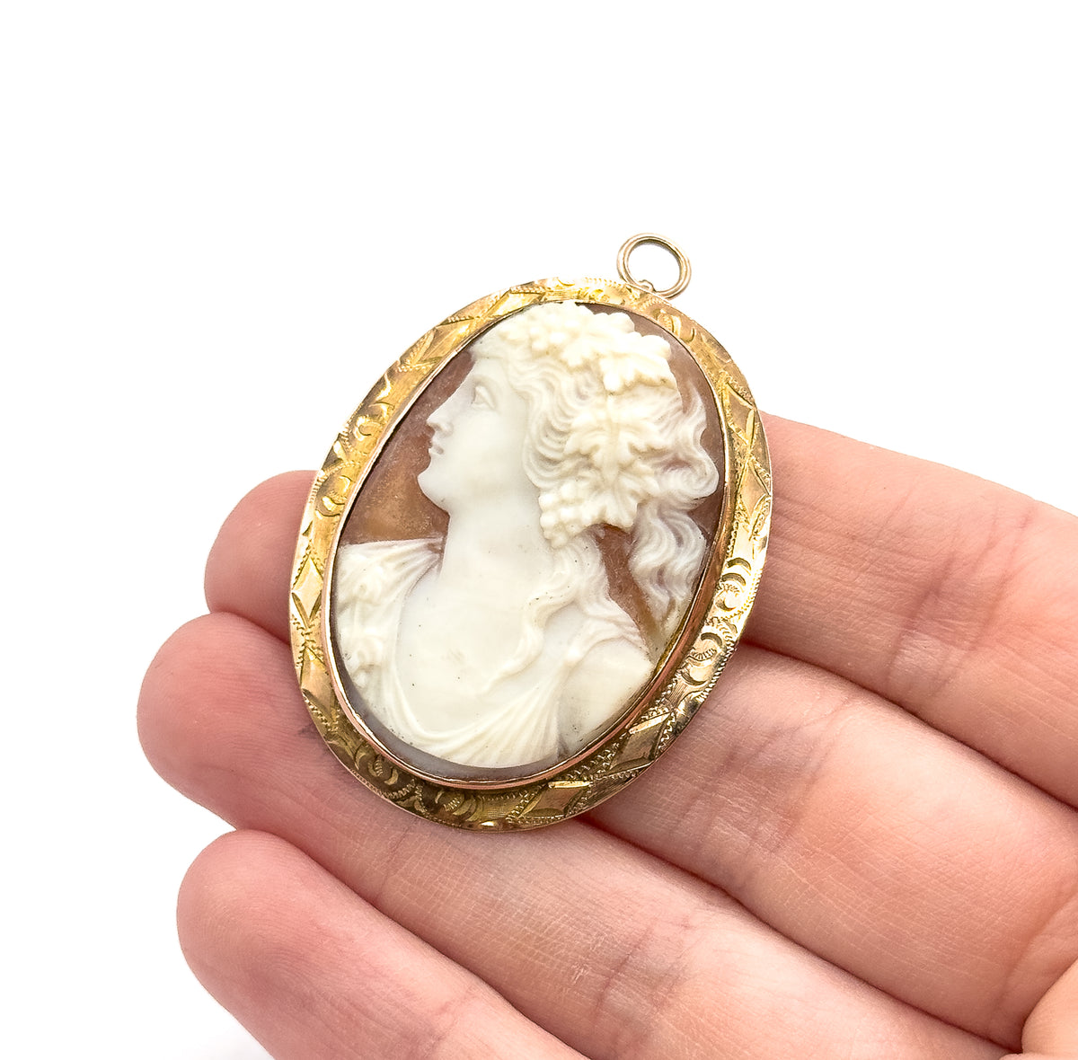 Victorian carved shell cameo brooch in 10K yellow gold, the creamy white Bacchante on warm tan within an engraved oval bezel with bail at top, shown resting on fingers for scale.