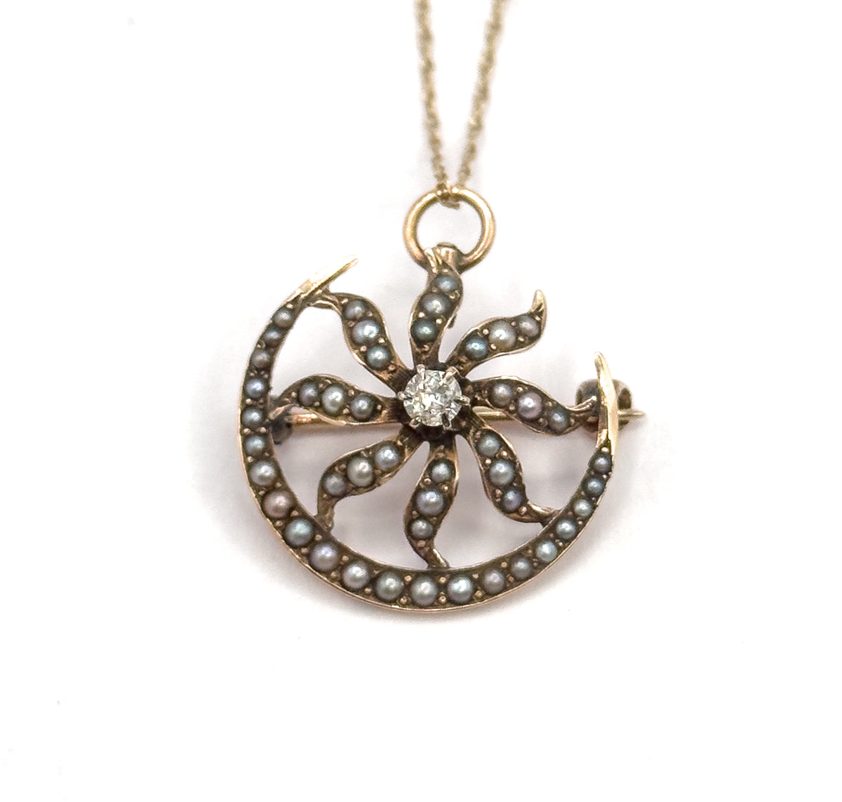 Straight-on front view with the chain rising vertically from the ring bail, the pearl-set sunburst arms curling around the central diamond and the pin stem projecting past the crescent at the right.