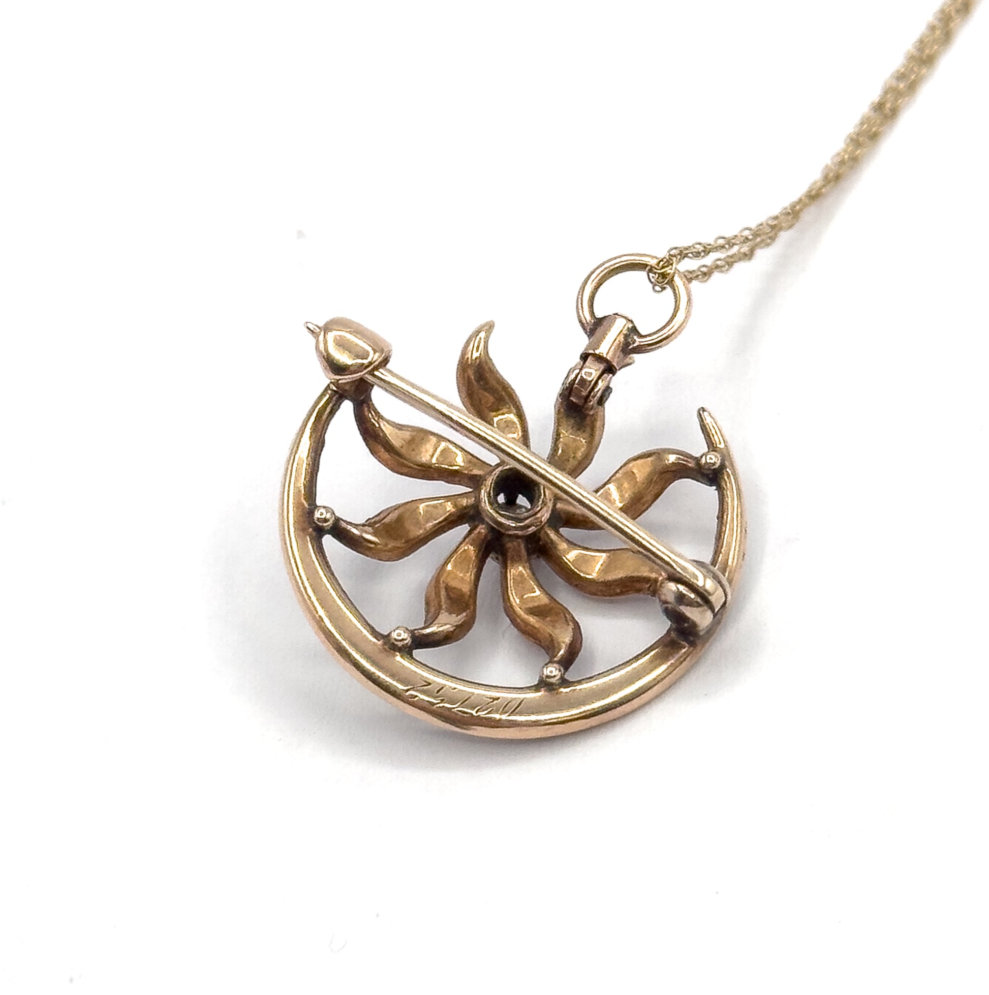 Angled back view showing the pin stem crossing the open sunburst, the collet holding the centre stone from behind, engraved script along the crescent, and the ring bail linking to the chain.