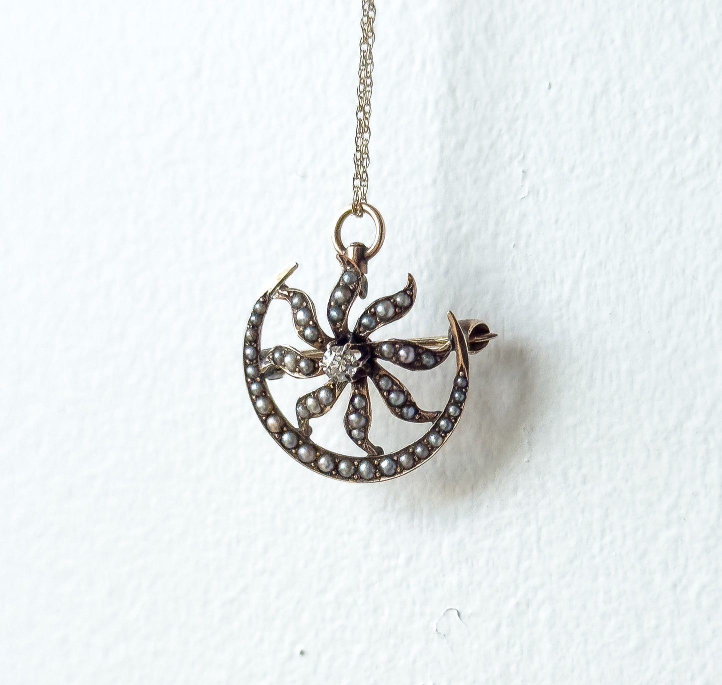 Front view hanging from its chain against a white wall, the crescent's seed pearl border and the pearl-set rays spiralling out from the central diamond seen face-on.
