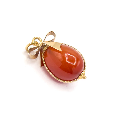 Diagonal view on white with the egg tipped to the lower right, showing the smooth gold cap over the top of the carnelian, the bow's loops and jump ring at upper left, and the bead at the tip.