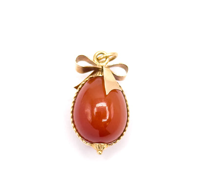 Mid-century carnelian egg charm in 14K yellow gold, shown front-on against a plain white background, the polished egg framed by twisted cable wire with a ribbon bow and jump ring above.