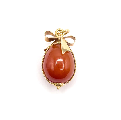 Closer front view filling the frame, the yellow gold bow's folded loops and pointed ribbon tail lying across the top of the carnelian egg, with the jump ring standing above.