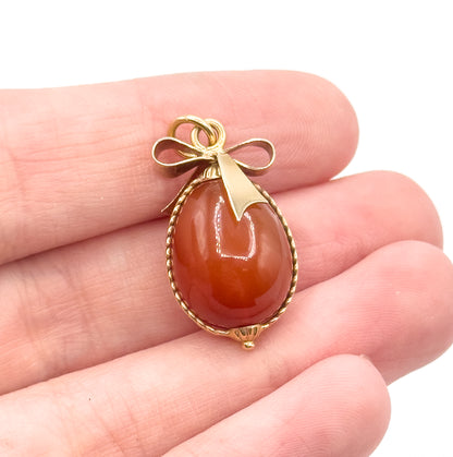 Held across three fingers for scale, the whole charm sits shorter than a fingertip is wide, the carnelian egg and its gold bow angled slightly toward the camera.