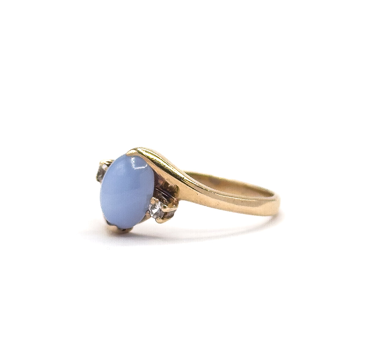 Angled side view with the ring upright on white, the periwinkle cabochon facing left in its four prongs and one colourless spinel catching light where the shank sweeps up.