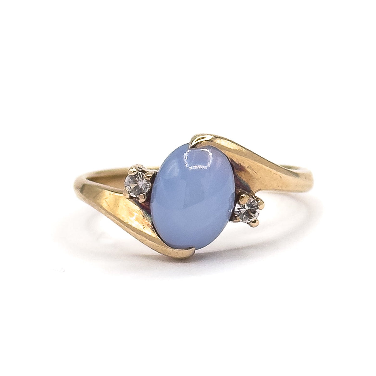 Face-on from the front, the oval periwinkle cabochon sits in its four prongs with a small colourless spinel flanking each side, the yellow gold shoulders sweeping past one another.