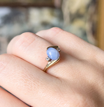 Midcentury lab-created sapphire cabochon and spinel ring in 10K yellow gold, shown on a finger, the periwinkle oval held in four prongs between two small colourless spinels.
