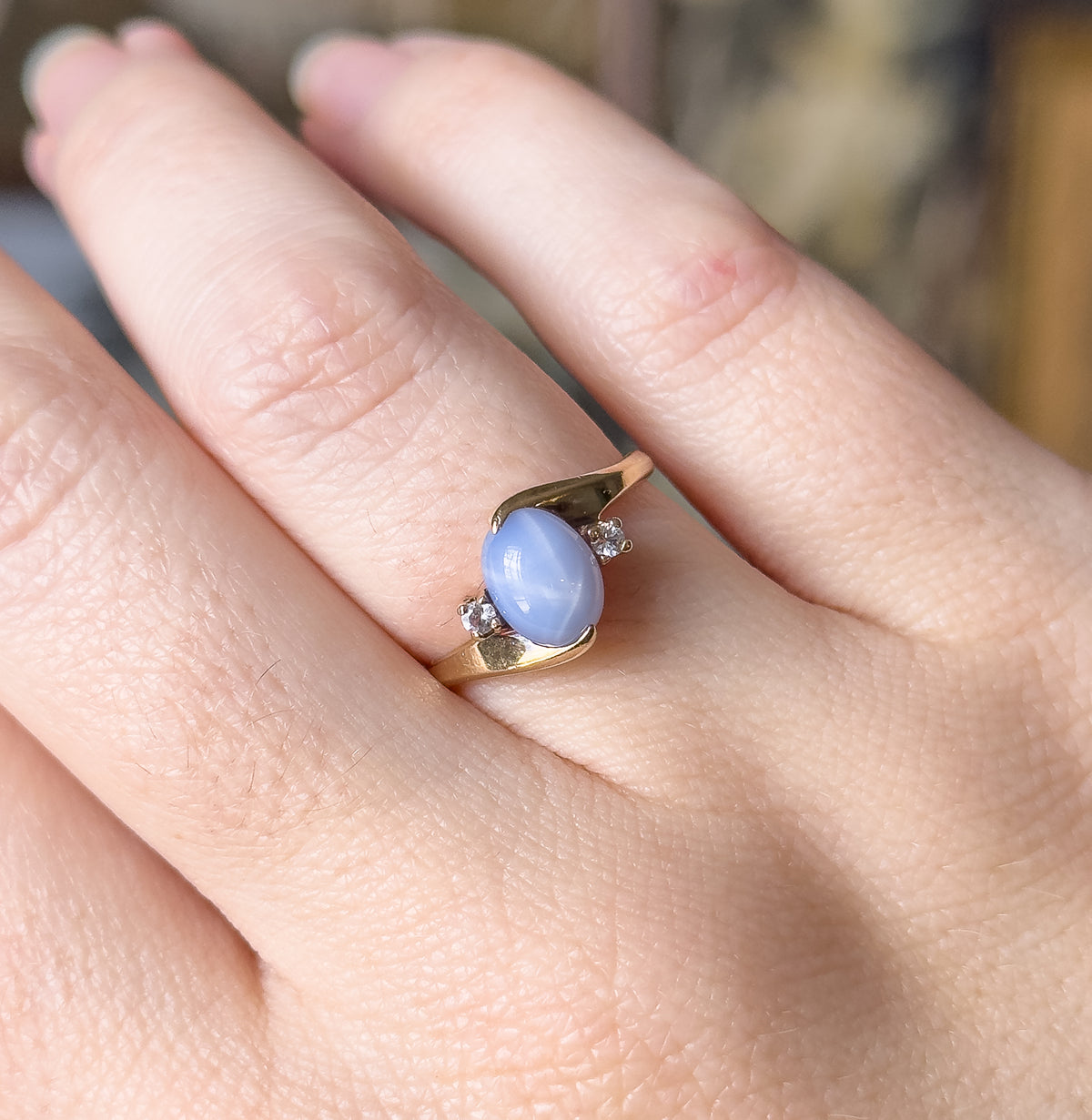 Angled close view on the hand, showing the swept yellow gold mounting curving past the domed periwinkle stone and tapering into the narrow shank.