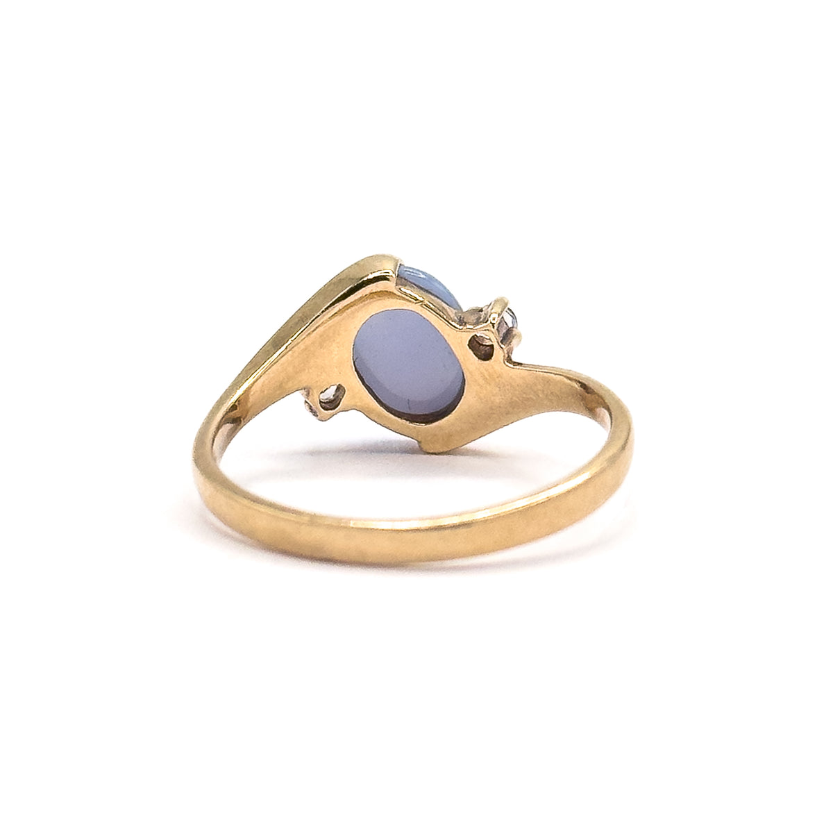 Viewed from behind and above, the open-backed mount frames the flat oval underside of the periwinkle cabochon, with a small colourless spinel in prongs to each side of the tapering shank.