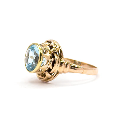 Angled from the front-left, the round light Swiss blue topaz faces the camera in its yellow gold bezel while the rose gold knotwork gallery and tapering shank recede to the right.