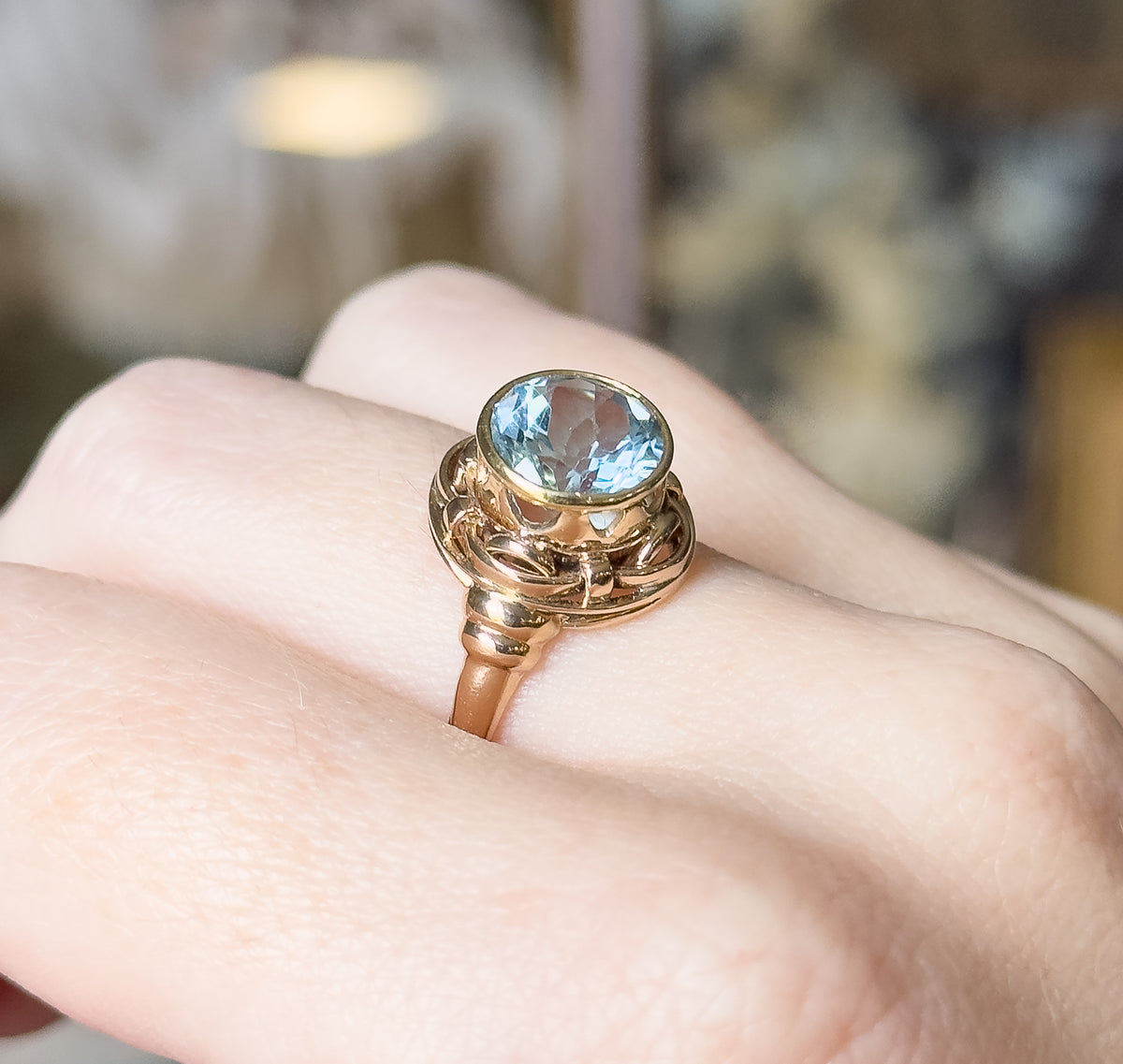 Angled view shown on a finger, the bezel-set topaz tilted toward the camera so the rose gold knotwork gallery and the tapering shank rising from the finger are visible in profile.