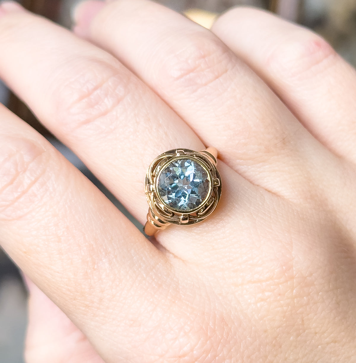Shown on the hand from above with the fingers together, the ring sitting flat so the round blue topaz and the looping knotwork frame are seen face-on against the skin for scale.