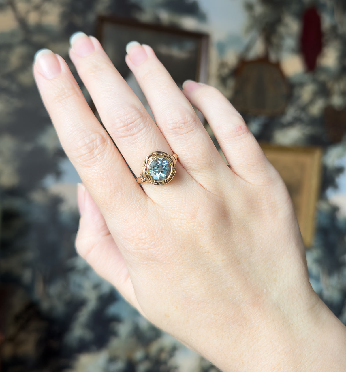 Fuller view of the raised hand and wrist, the ring small on the ring finger against a blurred backdrop, giving a sense of its scale on the whole hand.