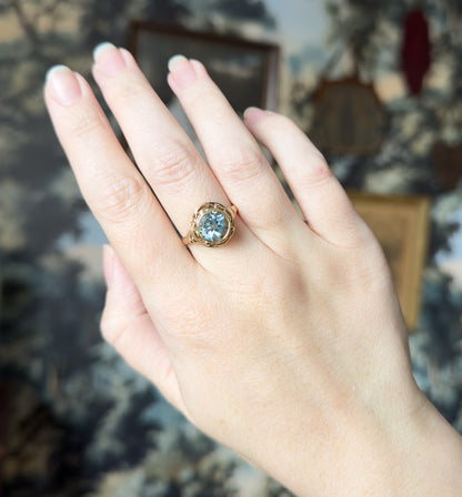 Fuller view of the raised hand and wrist, the ring small on the ring finger against a blurred backdrop, giving a sense of its scale on the whole hand.