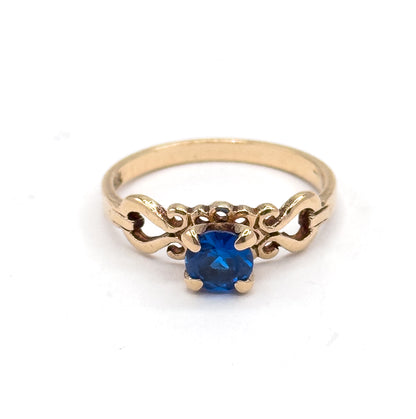Front view looking down into the four prongs, the round cobalt spinel centred between matching scrolled openwork shoulders with the full circle of the yellow gold shank rising behind.