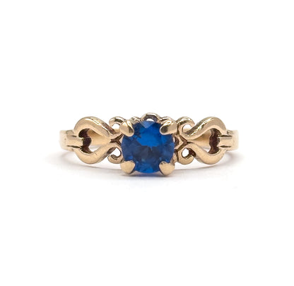 Eye-level front view with the ring standing upright, the round cobalt spinel facing the camera in its four prongs and the S-scroll shoulders flanking it as the yellow gold shank drops out of frame.