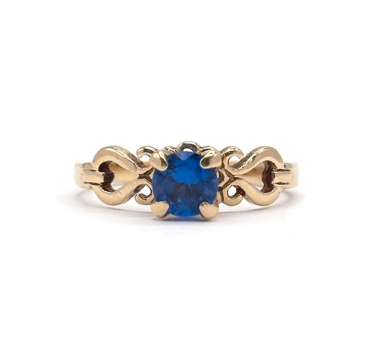 Eye-level front view with the ring standing upright, the round cobalt spinel facing the camera in its four prongs and the S-scroll shoulders flanking it as the yellow gold shank drops out of frame.