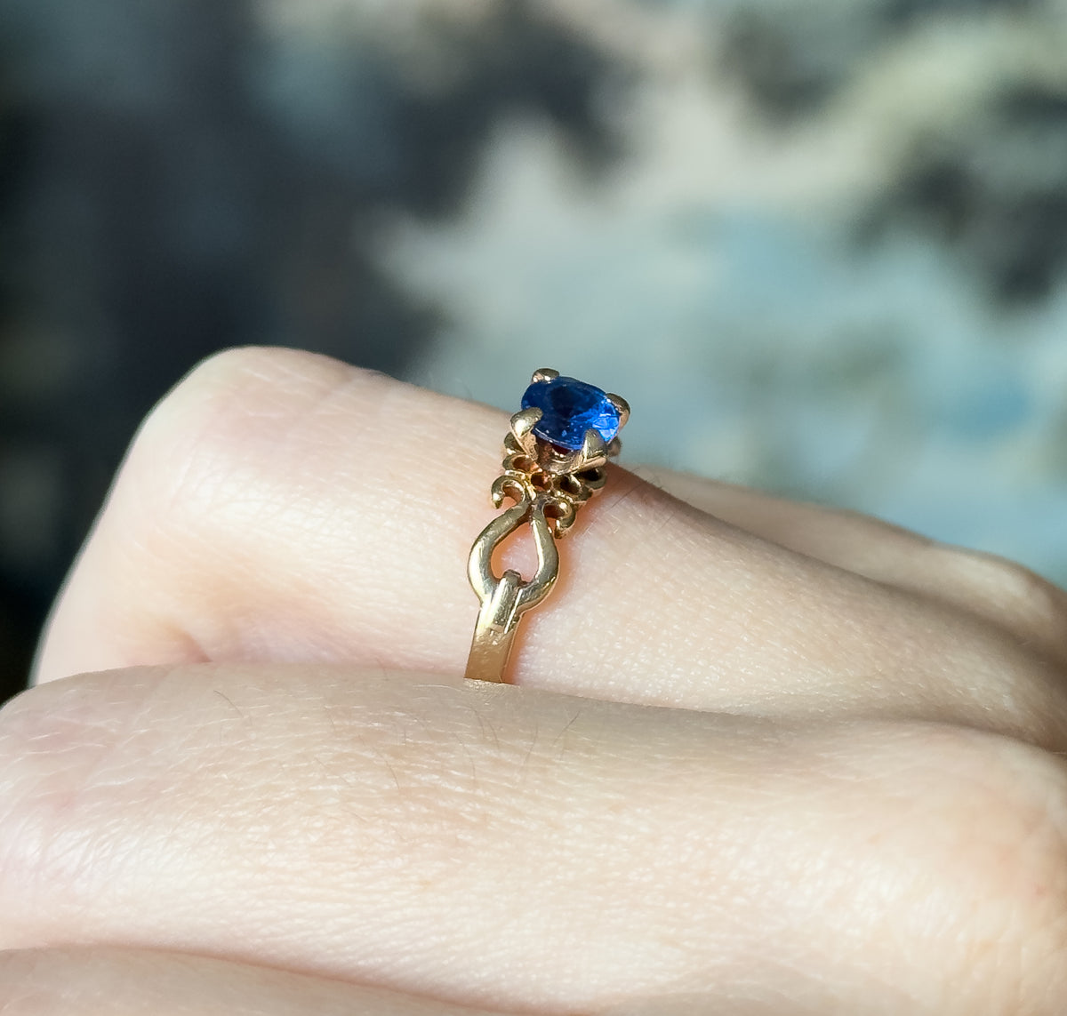 Mid-century lab-created blue spinel ring in 10K yellow gold, shown on a finger in close-up, the round cobalt stone held by four prongs above a scrolled, openwork shank.