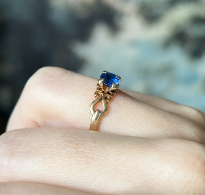 Mid-century lab-created blue spinel ring in 10K yellow gold, shown on a finger in close-up, the round cobalt stone held by four prongs above a scrolled, openwork shank.