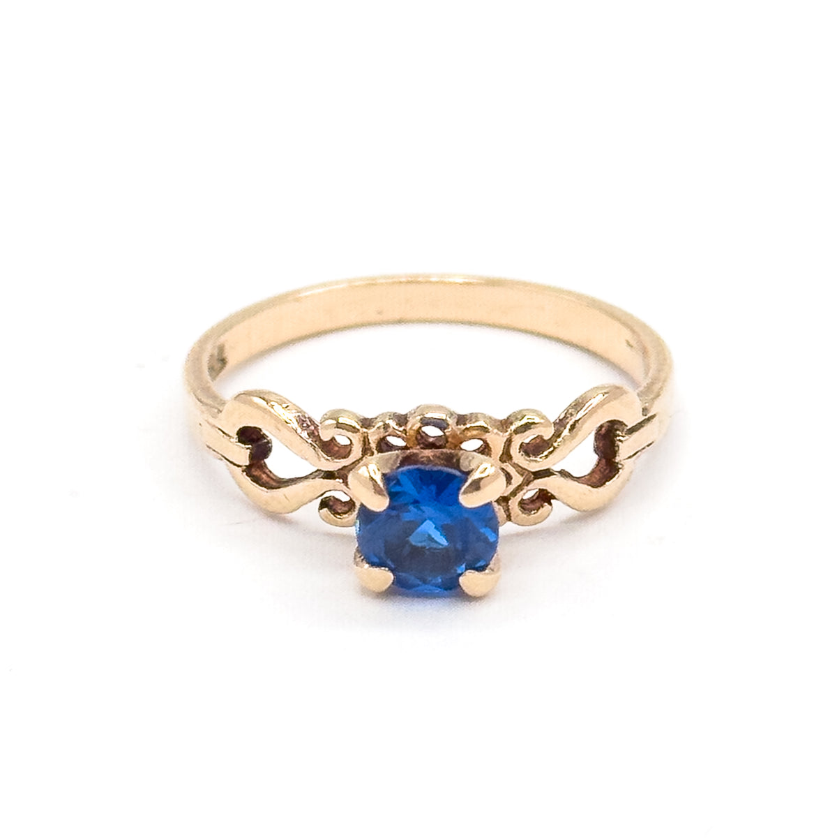 Standing upright on a plain white background, the ring faces the camera so the round cobalt spinel, its four prongs and the symmetrical scrollwork either side are fully visible.
