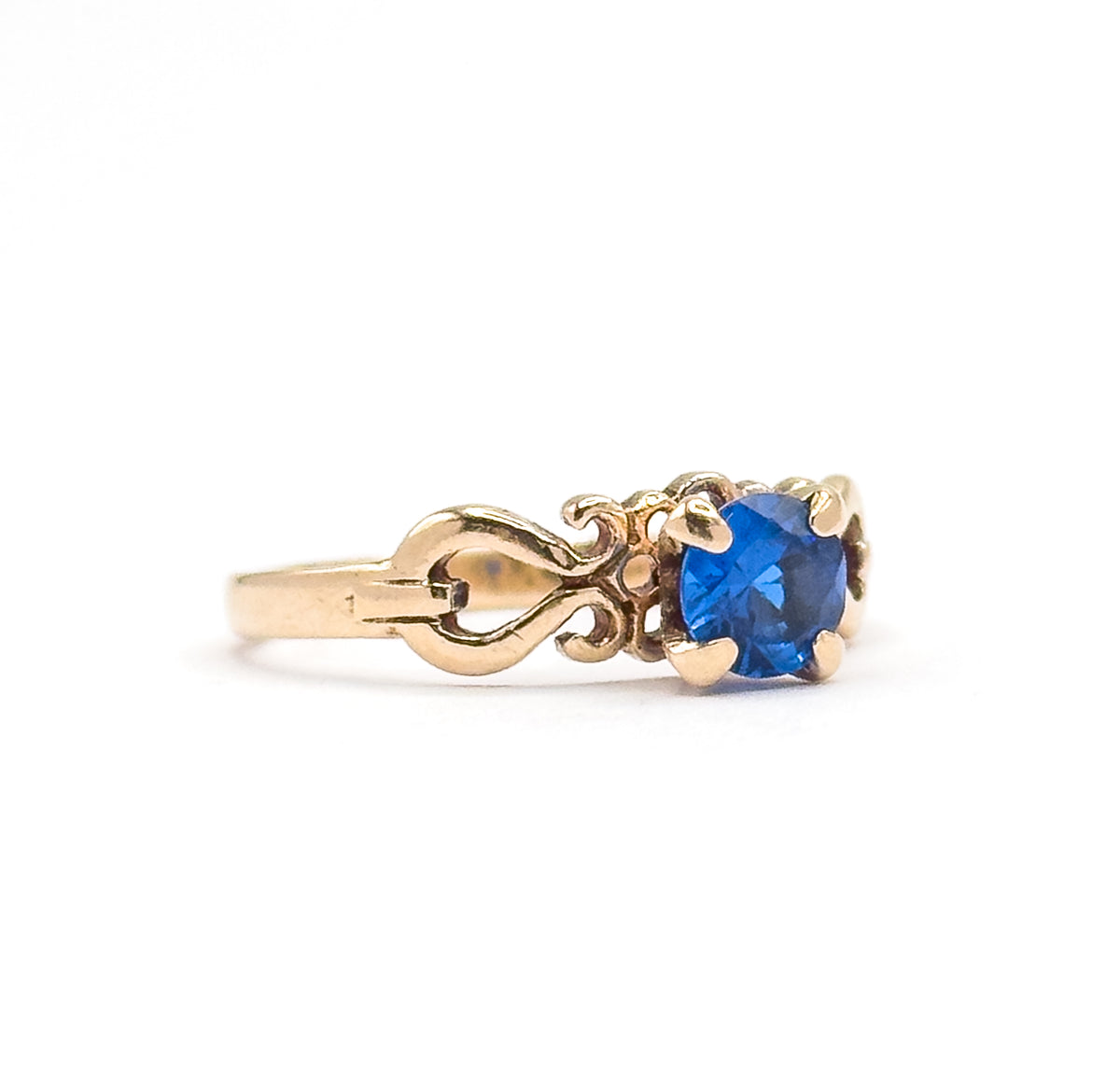 Profile view from the side on white, the round cobalt spinel seen edge-on in its four prongs above the looping scrolled shoulder and plain yellow gold shank.