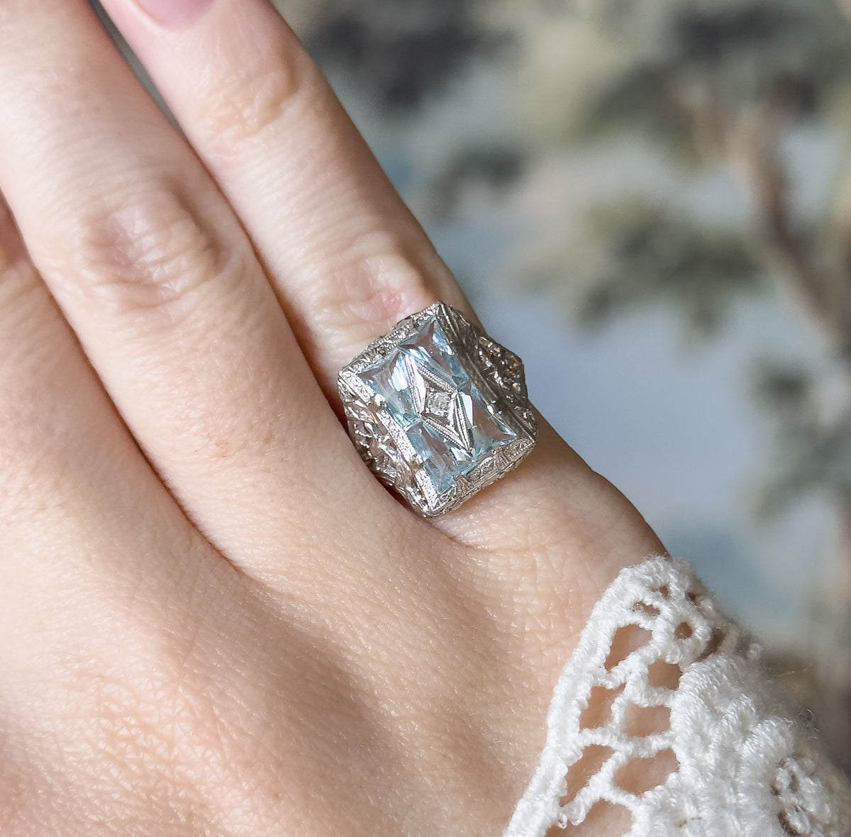 An Art Deco aquamarine and diamond filigree ring in 14K white gold, shown on a finger, its square filigree face set with four icy blue baguette stones around a small central diamond.