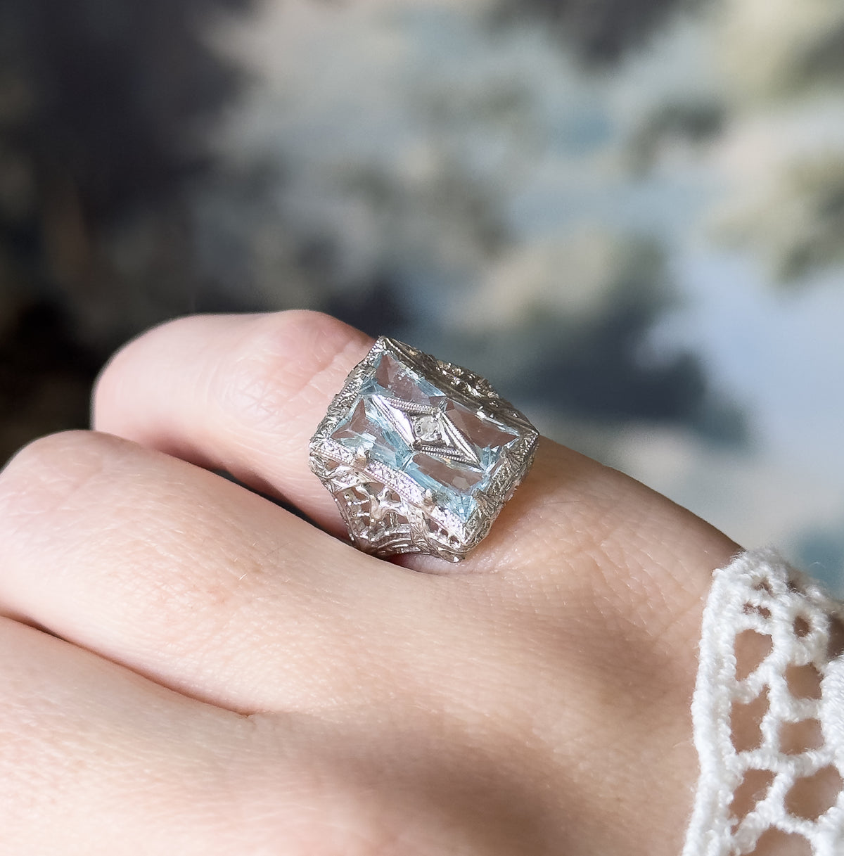 Angled three-quarter view shown on a finger, the square face tilted so the four aquamarines and central diamond catch light against a soft blue-green backdrop, with a lace cuff at the wrist.