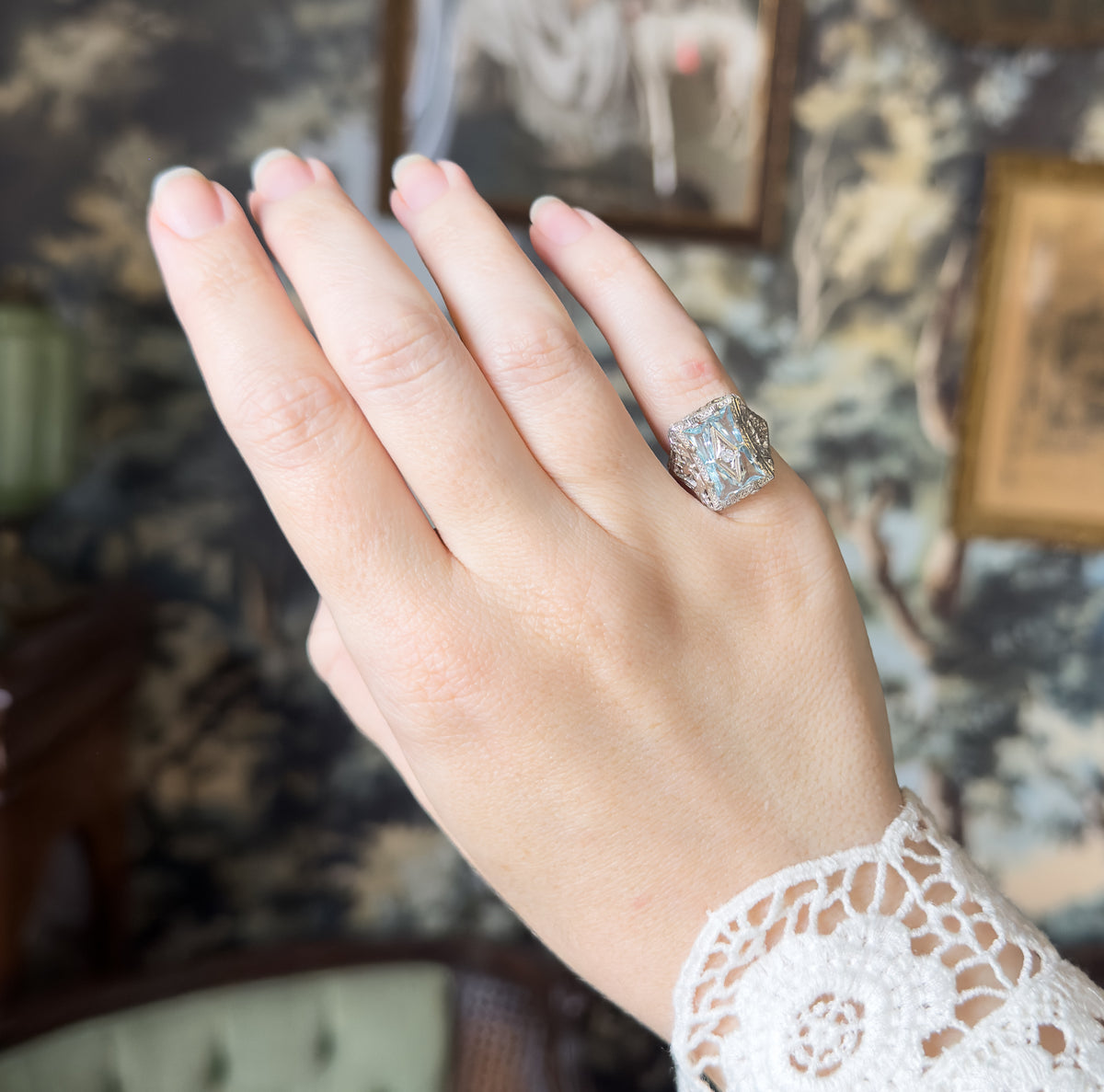 The whole hand raised with fingers together, the ring small on the ring finger, giving a sense of its scale against framed pictures on the patterned wall behind.